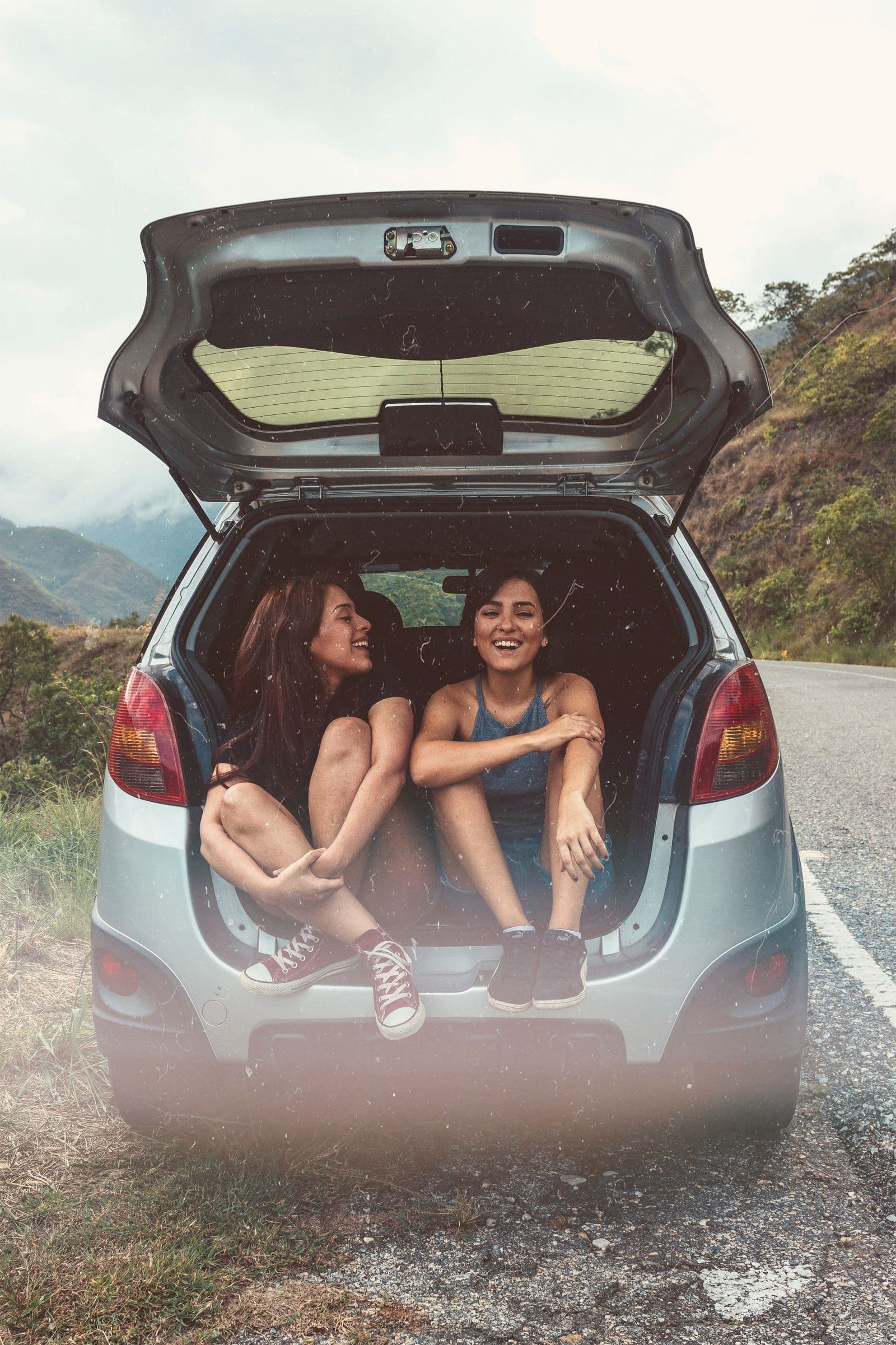 Two friends sitting in the open boot of a car on a mountain road