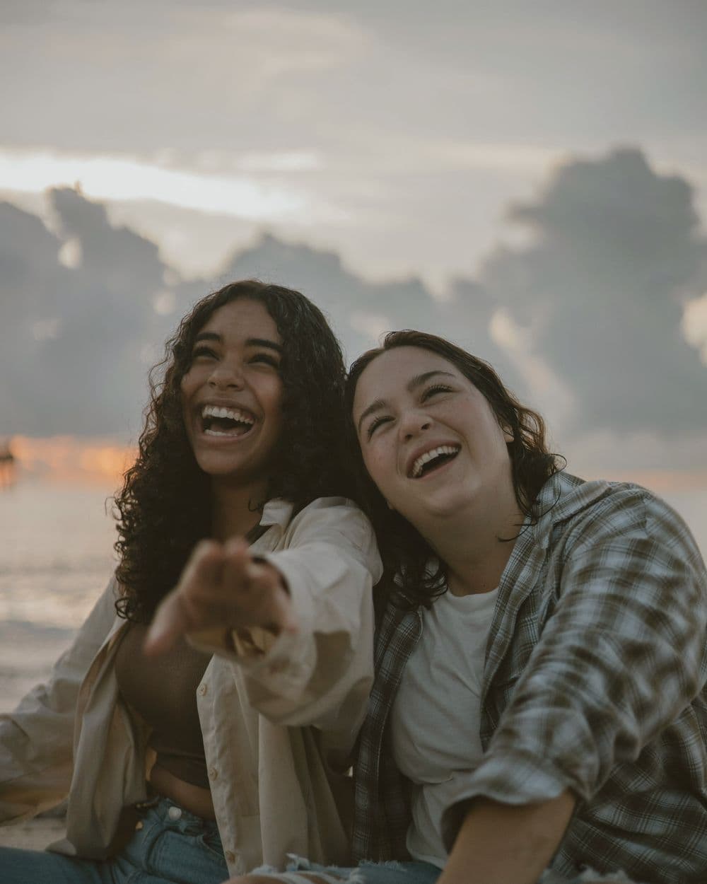 Two friends sitting together under a cloudy sky, one pointing, both laughing