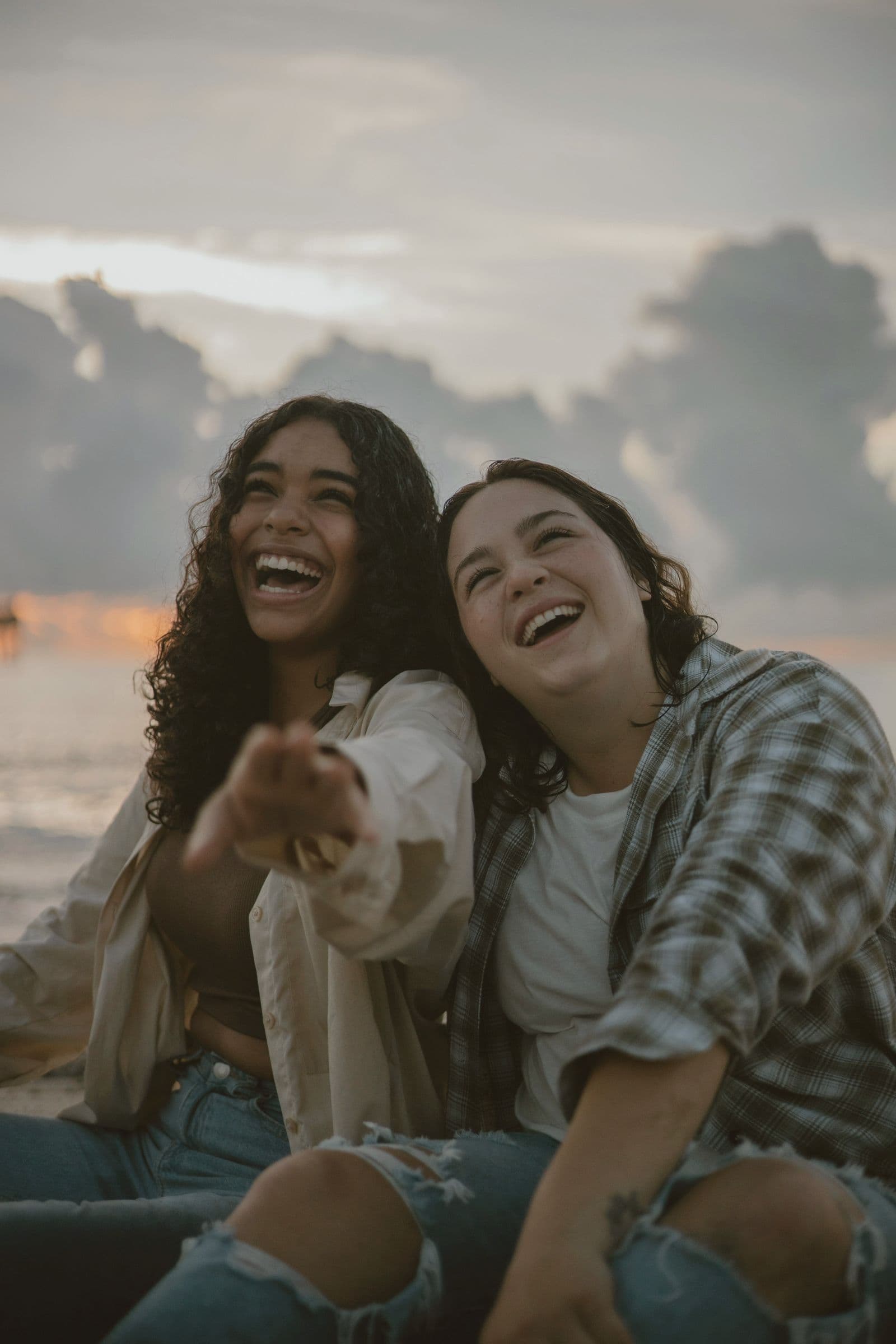 Two friends sitting together under a cloudy sky, one pointing, both laughing